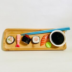 May include: A bamboo serving tray with sushi rolls, nigiri, and a small bowl of soy sauce. Blue chopsticks rest on the tray. The sushi includes a shrimp, salmon, and tuna roll, with a side of wasabi.