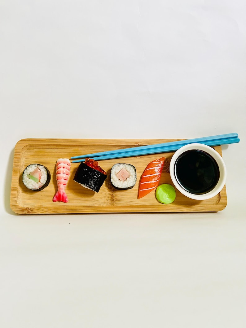 May include: A bamboo serving tray with sushi rolls, nigiri, and a small bowl of soy sauce. Blue chopsticks rest on the tray. The sushi includes a shrimp, salmon, and tuna roll, with a side of wasabi.
