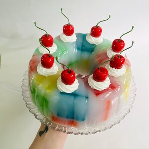 May include: A multi-coloured jelly dessert, resembling a rainbow, adorned with whipped cream and vibrant red cherries. The jelly is presented on a clear, decorative glass plate, creating a visually appealing treat.