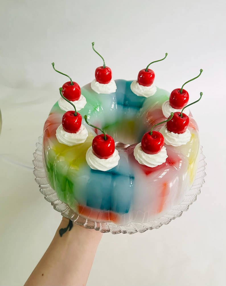 May include: A multi-coloured jelly dessert, resembling a rainbow, adorned with whipped cream and vibrant red cherries. The jelly is presented on a clear, decorative glass plate, creating a visually appealing treat.