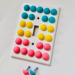 May include: A white light switch cover with a light switch in the centre. The cover is decorated with 30 colourful round buttons in three rows of ten. The top row is teal, the middle row is yellow, and the bottom row is pink. Two extra buttons are shown below the cover, one teal and one pink.