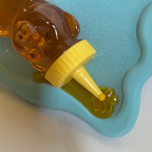 May include: A yellow squeeze bottle with a clear, honey-colored liquid is being squeezed onto a blue silicone mold. The liquid is forming a flower shape in the mold.