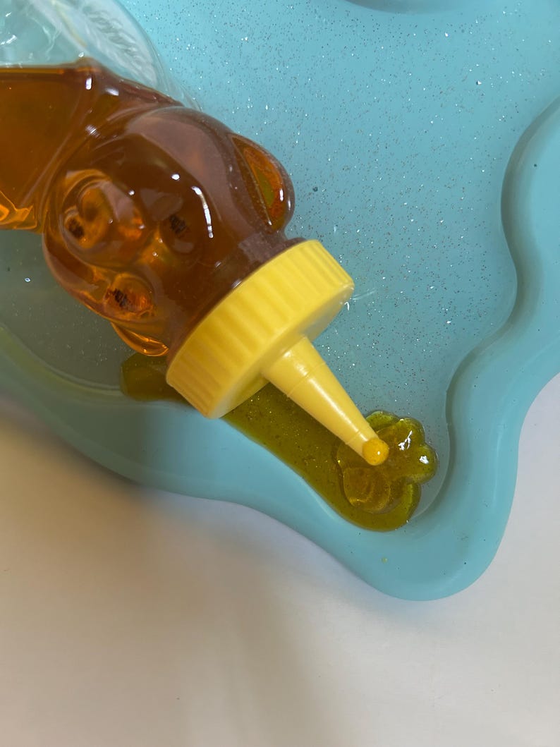 May include: A yellow squeeze bottle with a clear, honey-colored liquid is being squeezed onto a blue silicone mold. The liquid is forming a flower shape in the mold.