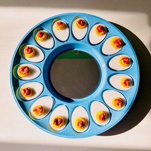 Puede incluir: Un porta huevos azul y blanco con 12 compartimentos individuales, cada uno con un huevo relleno con yema amarilla y pimentón rojo.