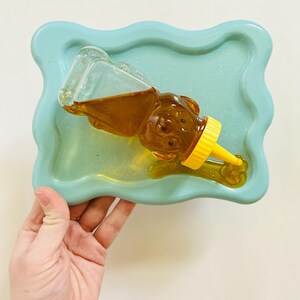 May include: A light blue resin tray with a wavy edge, holding a clear glass honey bear bottle with a yellow lid and spout. The honey is a golden color.