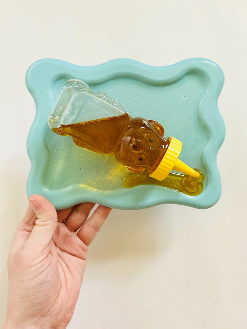 May include: A light blue resin tray with a wavy edge, holding a clear glass honey bear bottle with a yellow lid and spout. The honey is a golden color.