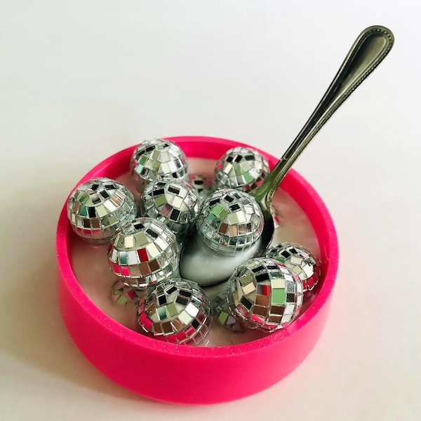 May include: A vibrant pink bowl filled with miniature disco balls and a silver spoon. The disco balls are covered in reflective mirrored tiles, creating a dazzling effect. The spoon rests in a creamy, light-colored substance.