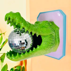 Pode incluir: Decora&ccedil;&atilde;o de parede com cabe&ccedil;a de jacar&eacute; verde segurando uma bola de discoteca prateada. O jacar&eacute; &eacute; montado em uma moldura retangular lil&aacute;s claro e azul. A boca do jacar&eacute; est&aacute; aberta e a bola de discoteca est&aacute; dentro.