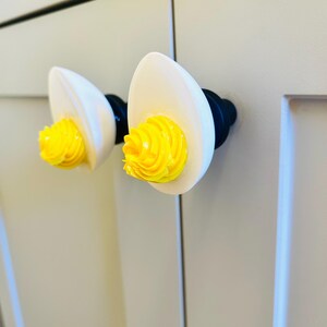 May include: Two white ceramic cabinet knobs shaped like eggs with yellow swirls on top. The knobs are mounted on a white cabinet door.