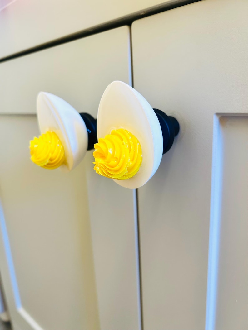May include: Two white ceramic cabinet knobs shaped like eggs with yellow swirls on top. The knobs are mounted on a white cabinet door.
