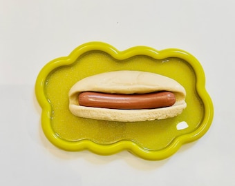 May include: A yellow cloud-shaped dish with a hot dog inside. The hot dog is in a white bun and has a brown sausage.