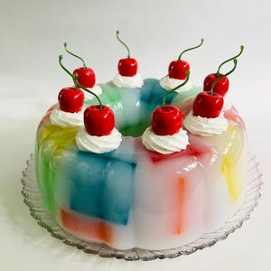 May include: A multi-coloured jelly dessert on a clear glass plate. The jelly is topped with whipped cream and bright red cherries. The dessert has a retro, vintage aesthetic.