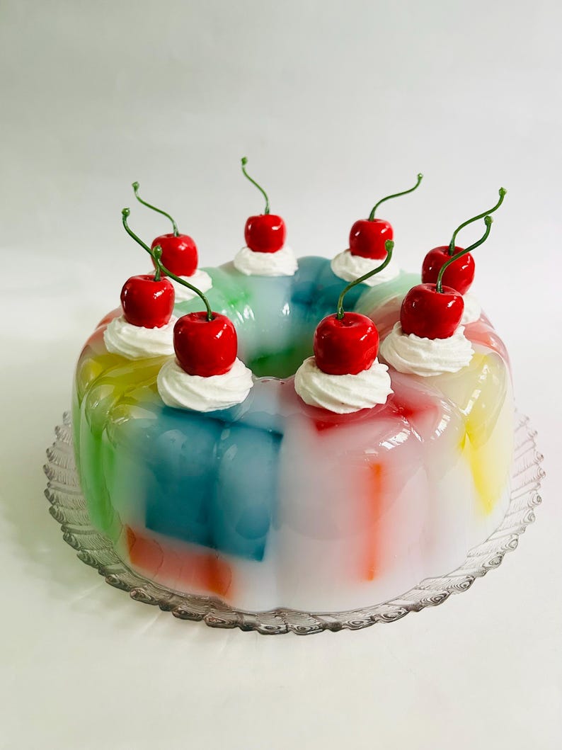 May include: A multi-coloured jelly dessert on a clear glass plate. The jelly is topped with whipped cream and bright red cherries. The dessert has a retro, vintage aesthetic.