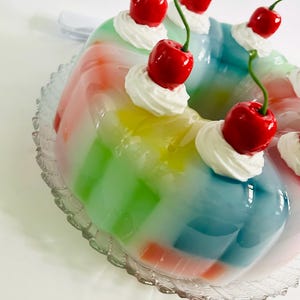 May include: A vibrant, multi-coloured jelly dessert, ring-shaped, with layers of red, orange, yellow, green, and blue. Each section is topped with a swirl of white cream and a bright red cherry. It rests on a clear, decorative glass plate.