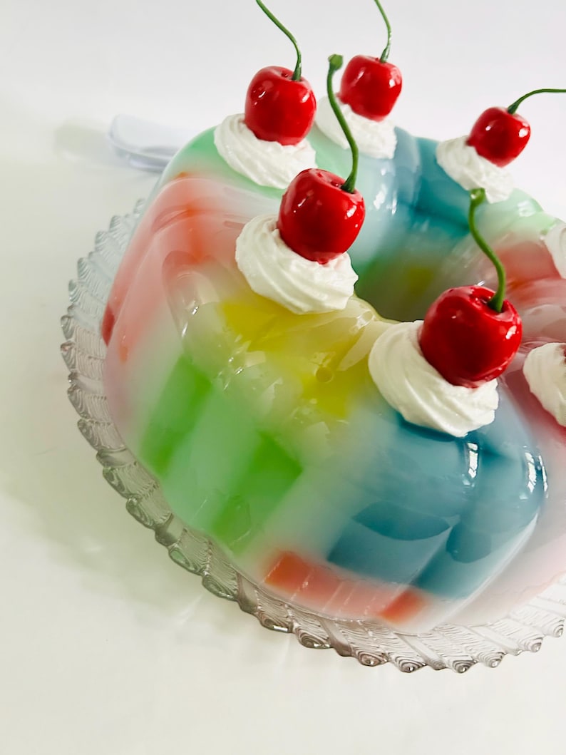 May include: A vibrant, multi-coloured jelly dessert, ring-shaped, with layers of red, orange, yellow, green, and blue. Each section is topped with a swirl of white cream and a bright red cherry. It rests on a clear, decorative glass plate.
