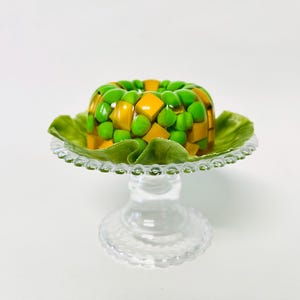 Puede incluir: Un bol de vidrio transparente lleno de un postre de gelatina amarillo y verde, decorado con guisantes verdes. El bol está sentado sobre un pedestal de vidrio transparente con un borde festoneado.