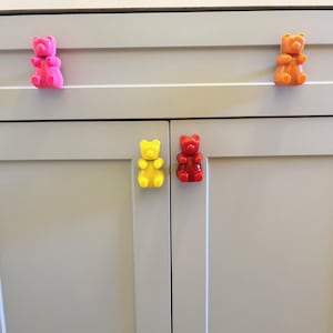 Op de afbeelding: Vier kastknopjes in de vorm van een gummybeer in verschillende kleuren: roze, oranje, geel en rood. De knoppen zijn bevestigd aan een witte kastdeur.