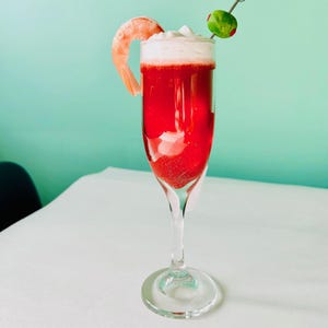May include: A red cocktail with a white foamy top in a tall glass with a shrimp garnish and a green olive on a cocktail stick. The drink is on a white tablecloth.