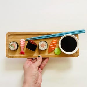 May include: A bamboo tray holds an assortment of sushi, including rolls, shrimp, and nigiri, with a small bowl of soy sauce and a pair of blue chopsticks. A small green ball of wasabi sits next to the soy sauce. The sushi is arranged on a light-colored wooden tray.