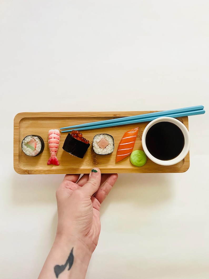 May include: A bamboo tray holds an assortment of sushi, including rolls, shrimp, and nigiri, with a small bowl of soy sauce and a pair of blue chopsticks. A small green ball of wasabi sits next to the soy sauce. The sushi is arranged on a light-colored wooden tray.