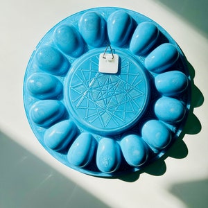 May include: A blue ceramic egg holder with a starburst design in the center. The holder has 12 individual egg-shaped compartments.