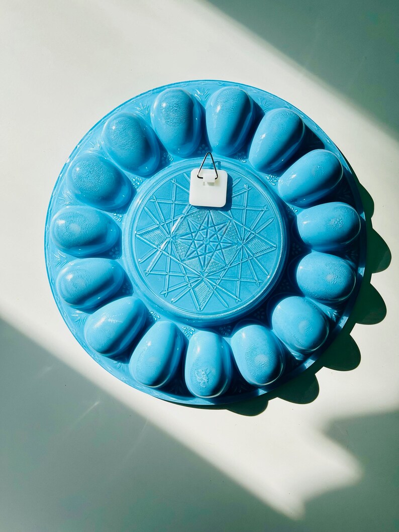 May include: A blue ceramic egg holder with a starburst design in the center. The holder has 12 individual egg-shaped compartments.