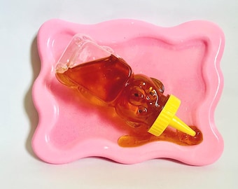 May include: A clear, bear-shaped honey bottle with a yellow cap, resting on a pink, wavy-edged tray. The bottle is filled with amber-colored honey, with some honey spilled onto the tray. The tray has a glossy finish.