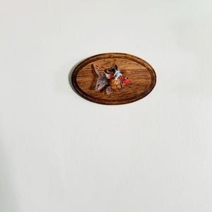 May include: A small, orange and white goldfish figurine mounted on a wooden plaque. The plaque is oval shaped and has a dark wood grain.
