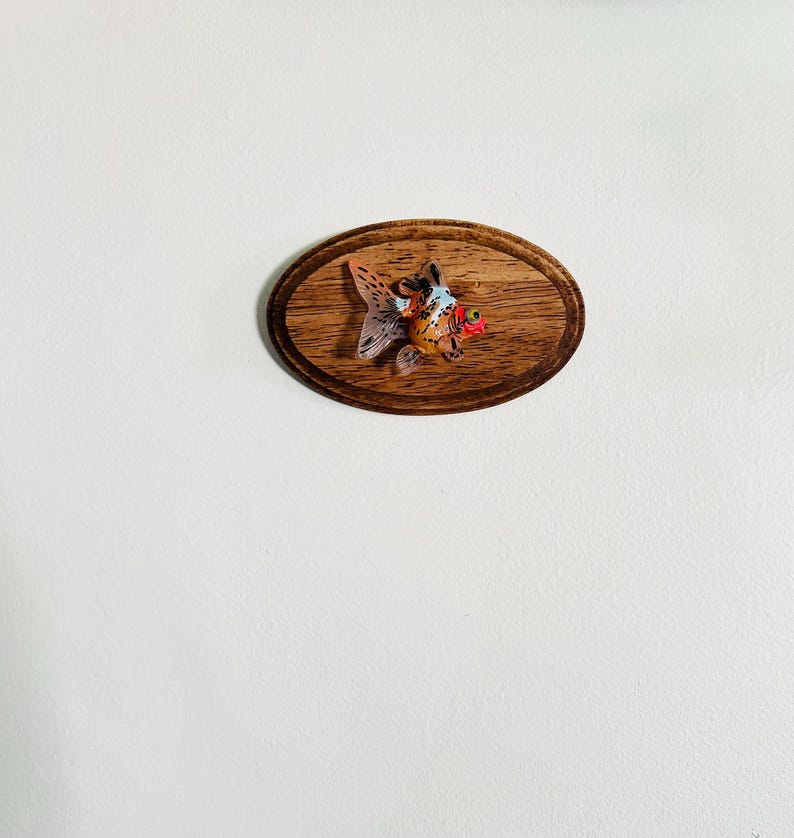 May include: A small, orange and white goldfish figurine mounted on a wooden plaque. The plaque is oval shaped and has a dark wood grain.