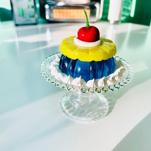 May include: A blue and yellow pineapple upside-down cake with a red cherry on top, displayed on a clear glass cake stand.
