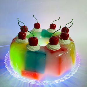 May include: A vibrant, layered dessert with a rainbow gradient effect. The dessert is topped with whipped cream and cherries. It sits on a clear, decorative plate.