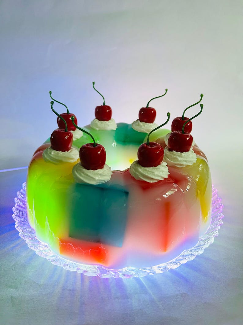 May include: A vibrant, layered dessert with a rainbow gradient effect. The dessert is topped with whipped cream and cherries. It sits on a clear, decorative plate.