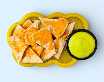 May include: Overhead shot of a yellow cloud-shaped serving dish filled with tortilla chips topped with orange cheese. A small black bowl of green dip sits next to the chips on a light blue background.