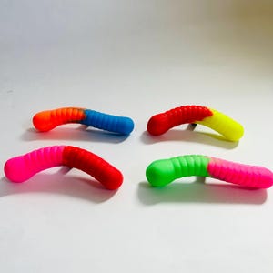 Puede incluir: Cuatro señuelos de plástico con forma de gusano, cada uno con una combinación de colores diferente. Los señuelos están dispuestos en fila sobre una superficie blanca.