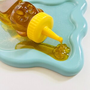 May include: A yellow bear-shaped honey dispenser with a clear glass body and a yellow spout pouring honey onto a light blue, wavy, ceramic dish.