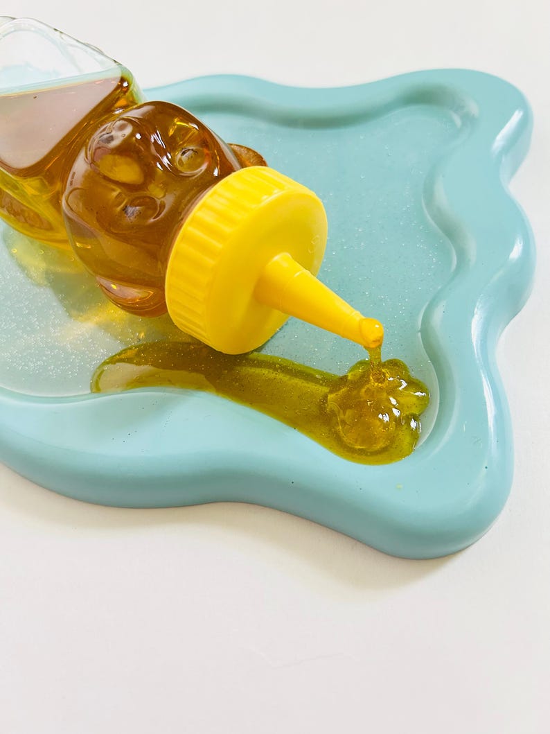 May include: A yellow bear-shaped honey dispenser with a clear glass body and a yellow spout pouring honey onto a light blue, wavy, ceramic dish.