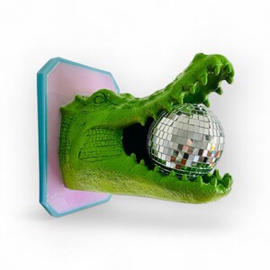 Pode incluir: Escultura de cabe&ccedil;a de jacar&eacute; verde segurando uma bola de discoteca prateada. A escultura &eacute; montada em uma base retangular rosa e azul. A boca do jacar&eacute; est&aacute; aberta e a bola de discoteca est&aacute; dentro.