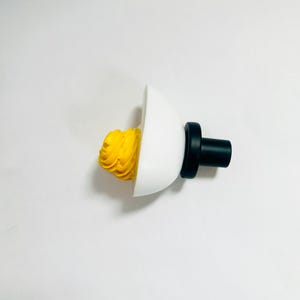 May include: A white ceramic egg-shaped bottle stopper with a black top and a yellow swirl of wax inside.