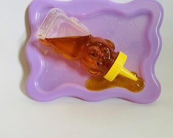May include: A clear plastic honey bear bottle with a yellow spout, tipped over on a purple tray with glitter. The honey has spilled out of the bottle and is pooling on the tray.
