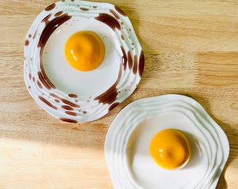 Puede incluir: Dos platos de cerámica blanca con forma de huevo frito. Los platos tienen diseños moteados en marrón y blanco. Cada plato tiene una yema de huevo de cerámica amarilla en el centro.