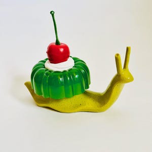 Könnte beinhalten: Eine grüne und gelbe Keramik-Schnecken-Skulptur mit einer grünen und weißen Geleeform auf dem Rücken. Die Geleeform ist mit einer roten Kirsche verziert.