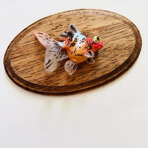 May include: A small, orange, white, and black goldfish figurine with a red mouth and a yellow eye. The figurine is sitting on a wooden oval-shaped base.