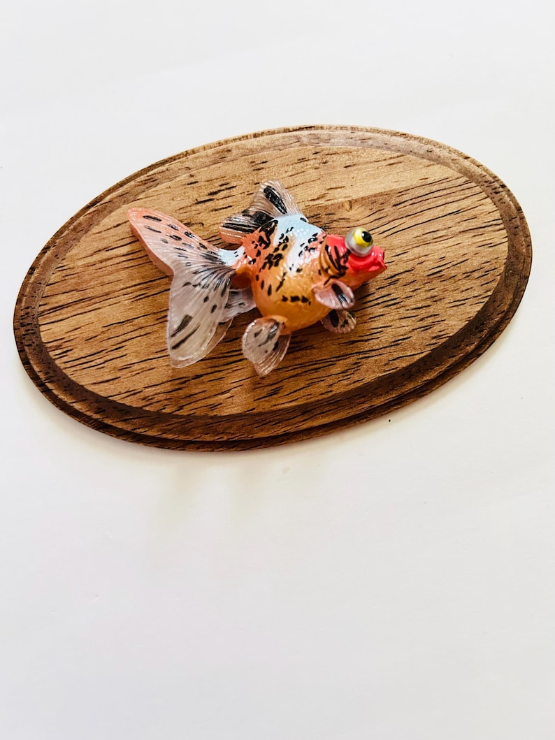 May include: A small, orange, white, and black goldfish figurine with a red mouth and a yellow eye. The figurine is sitting on a wooden oval-shaped base.