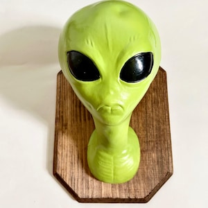 Puede incluir: Una escultura de cabeza de extraterrestre verde con ojos negros, montada en una placa de madera. El extraterrestre tiene una expresión seria y una barbilla ligeramente puntiaguda.