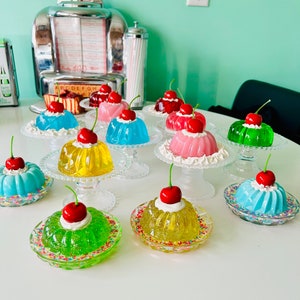 Puede incluir: Una mesa con una presentación de postres de gelatina de colores con cerezas rojas en la parte superior. Los postres están dispuestos en soportes para pasteles blancos con chispas en los platos.