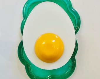 Puede incluir: Una pieza de arte de resina caprichosa con forma de huevo frito. El diseño presenta una yema amarilla brillante, un huevo blanco y un borde festoneado verde translúcido. La obra de arte es un artículo decorativo lúdico.