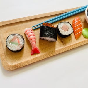 May include: A wooden serving tray holds an assortment of sushi rolls, including a shrimp nigiri, a salmon nigiri, and several maki rolls. Blue chopsticks rest on the tray, next to a small bowl of soy sauce and a green wasabi ball.