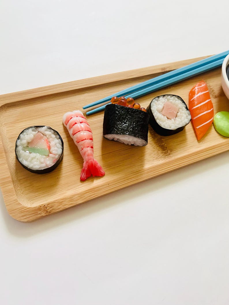 May include: A wooden serving tray holds an assortment of sushi rolls, including a shrimp nigiri, a salmon nigiri, and several maki rolls. Blue chopsticks rest on the tray, next to a small bowl of soy sauce and a green wasabi ball.