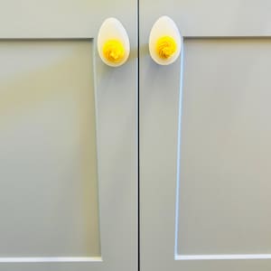 May include: Two white cabinet knobs shaped like eggs with yellow yolk details. The knobs are mounted on a light blue cabinet door.