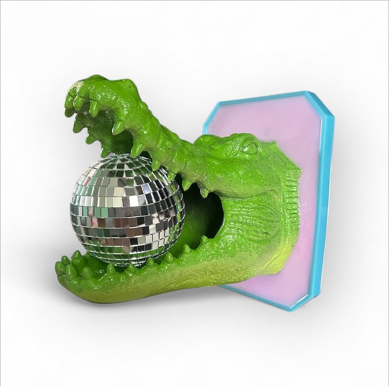 Pode incluir: Escultura de um jacar&eacute; verde segurando uma bola de discoteca prateada. O jacar&eacute; &eacute; montado em uma placa retangular rosa e azul. A escultura tem uma apar&ecirc;ncia texturizada e escamosa.